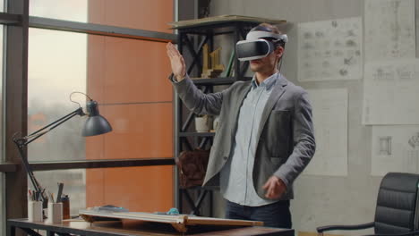 Modern-man-architect-in-the-office-with-large-windows-stands-in-a-virtual-reality-helmet-uses-gestures-to-manage-the-project-without-leaving-the-office.-Construction-control.-Design-project
